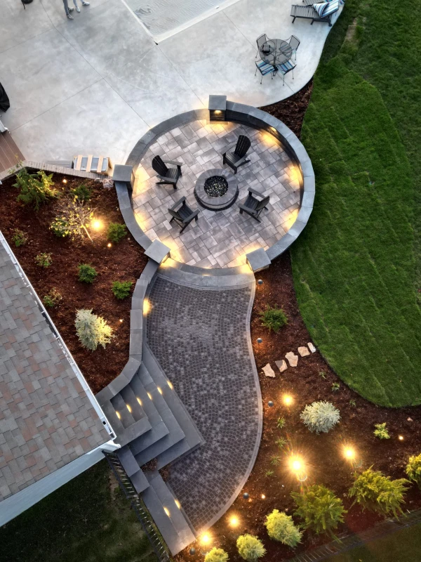 Aerial view of a modern backyard patio featuring a circular fire pit area with black Adirondack chairs, a curved paver walkway, outdoor lighting, landscaped greenery, and a mix of grass, mulch, and concrete surfaces.