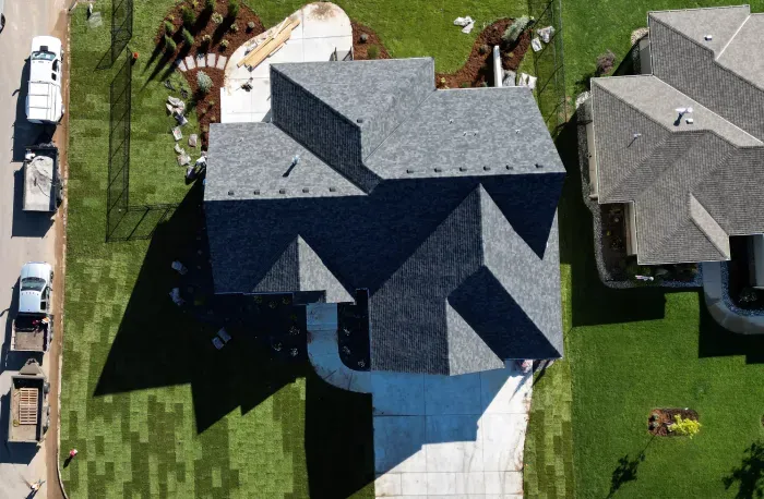 An aerial view of a house with a dark shingle roof, surrounded by a well-manicured lawn and landscaped yard. The driveway is visible, along with neatly arranged patches of sod indicating recent landscaping work. Nearby vehicles and landscaping materials are also present.