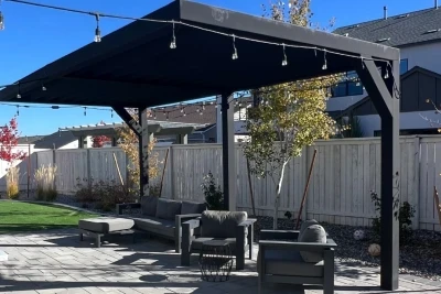 View of a shaded patio area with a modern pergola adorned with string lights, featuring comfortable outdoor furniture and surrounded by a landscaped yard, showcasing recent outdoor construction.