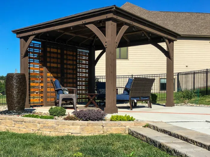 A cozy outdoor space featuring a wooden pergola with a lattice backdrop, complemented by comfortable seating and decorative plants, set against a grassy area and stone landscaping.