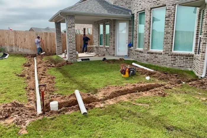 A backyard undergoing landscaping renovations, featuring freshly dug soil and laid-out irrigation pipes. Two workers are visible near the house, which has large windows and a covered porch, with a grassy area awaiting landscaping updates.