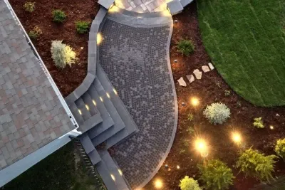 Aerial view of a beautifully constructed pathway with paver stones, illuminated by lights, winding through a landscaped garden with greenery and mulch, showcasing recent construction work.