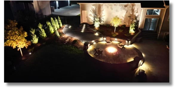 Landscape view of a beautifully lit backyard at night, featuring a circular fire pit surrounded by seating, illuminated pathways, and strategically placed plants and trees, creating a cozy outdoor space.