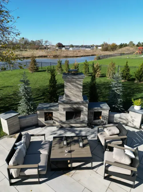 View of a cozy outdoor seating area featuring modern furniture arranged around a stone fireplace, set against a backdrop of a lush green lawn and a serene pond, showcasing recent construction work.