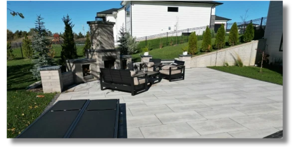A modern outdoor patio with a stylish seating area featuring black furniture. In the background, there is a stone fireplace and a landscaped yard with neatly trimmed trees and grass under a clear blue sky.