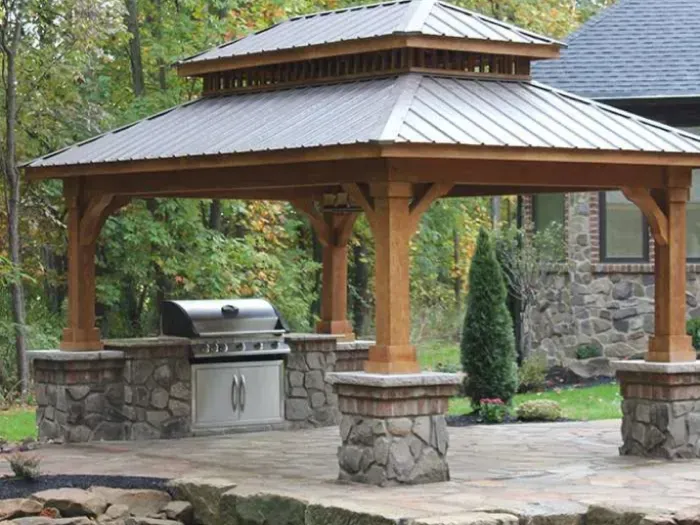 A spacious outdoor pergola featuring a stone base, providing shelter over a built-in grill area, surrounded by landscaped greenery and designed for cooking and entertaining.