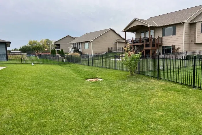 A grassy backyard bordered by a black fence, featuring a well-maintained lawn. In the background, two houses with decks are visible, along with some landscaping that includes shrubs and trees.