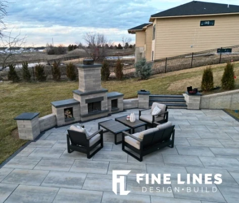 Landscape view of a beautifully designed outdoor patio featuring a modern seating area with gray furniture, a stone fireplace, and a well-maintained lawn in the background.