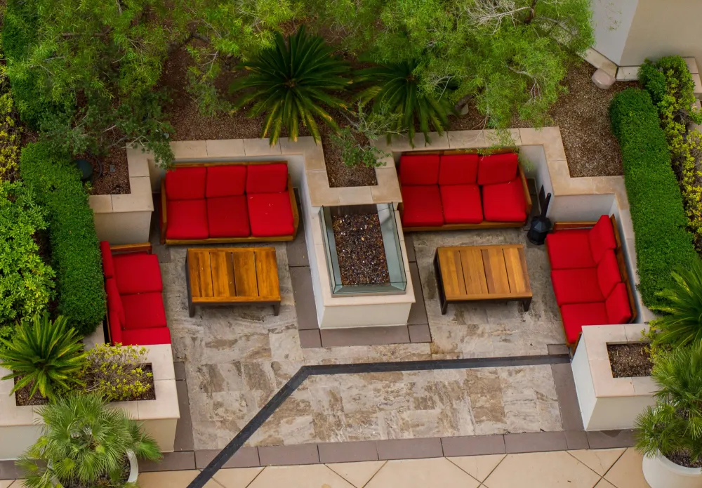 An aerial view of a cozy outdoor seating area featuring red cushioned chairs arranged around wooden tables, surrounded by neatly trimmed greenery and decorative planters.