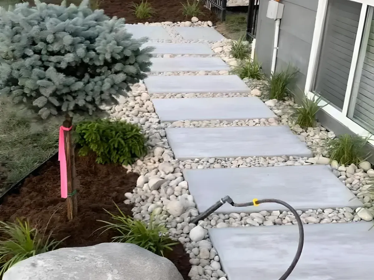 A landscaped path featuring large concrete stepping stones surrounded by decorative gravel, grass, and small plants, with a tree in the background and a garden hose resting nearby.