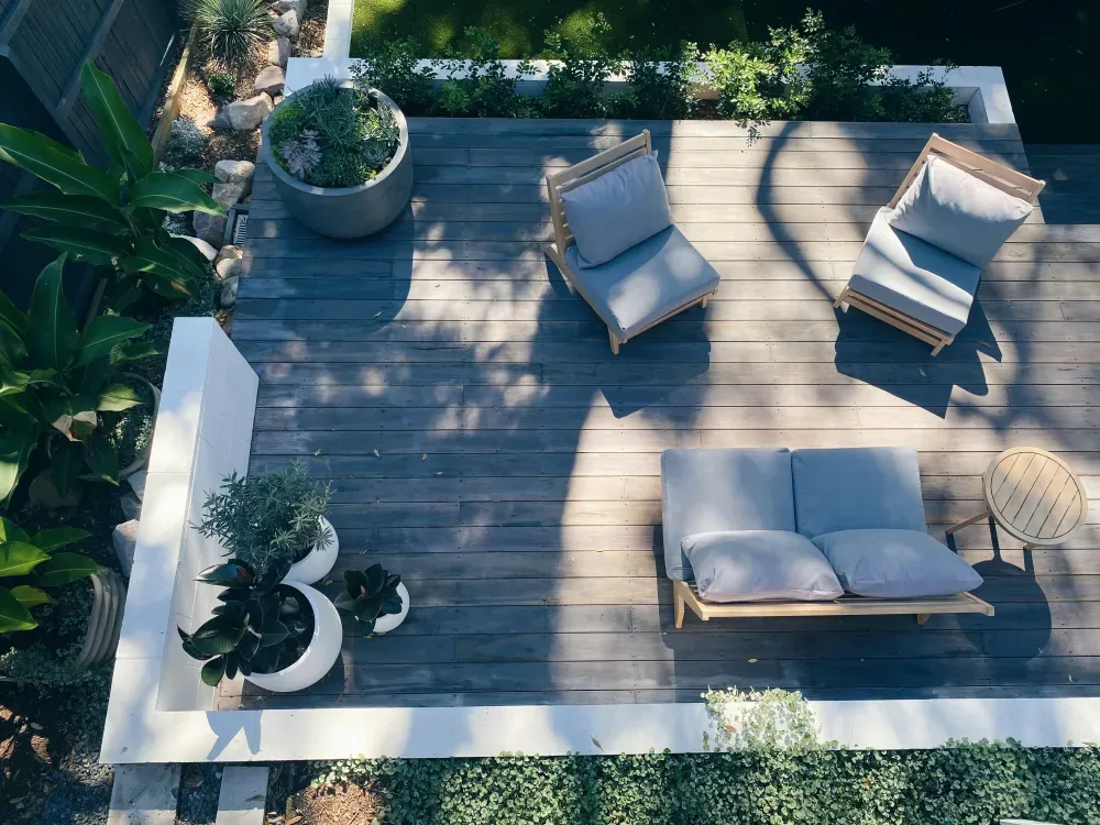 An overhead view of a stylish outdoor deck featuring wooden flooring, potted plants, and comfortable seating arrangements, including a couch and lounge chairs, surrounded by greenery.