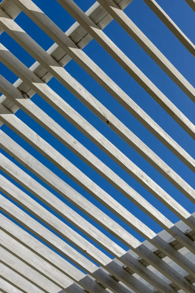 A close-up view of a wooden pergola structure, showcasing its slatted design against a clear blue sky, creating a play of light and shadow.